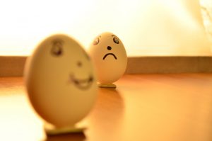 A sad expression drawn onto an egg that appears to be looking at another egg that appears happy